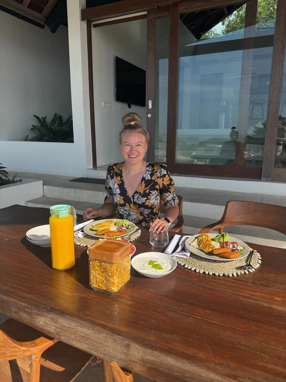 Annina sitzt lächelnd am langen Holztisch, vor ihr Orangensaft, Cornflakes, Joghurt und zwei Teller mit Frühstück