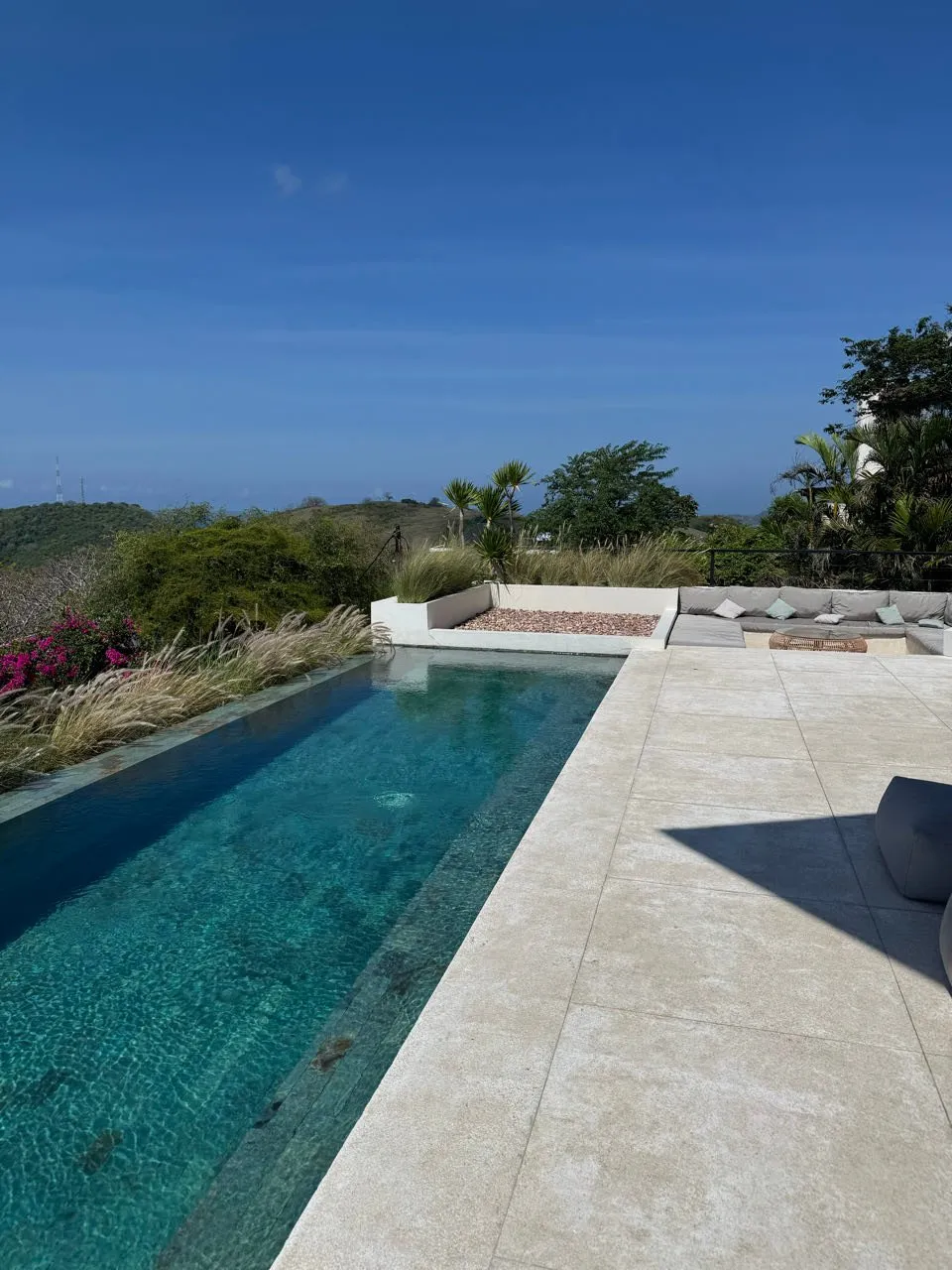 Langer, schmaler Pool mit hellen Steinplatten, dahinter eine Sitzecke mit grauen Kissen, pinke Blüten und Hügel unter blauem Himmel