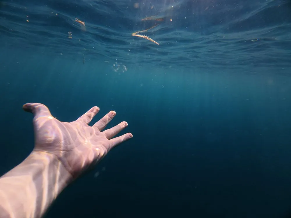 A hand reaches into the deep blue underwater, with the light of the surface above it