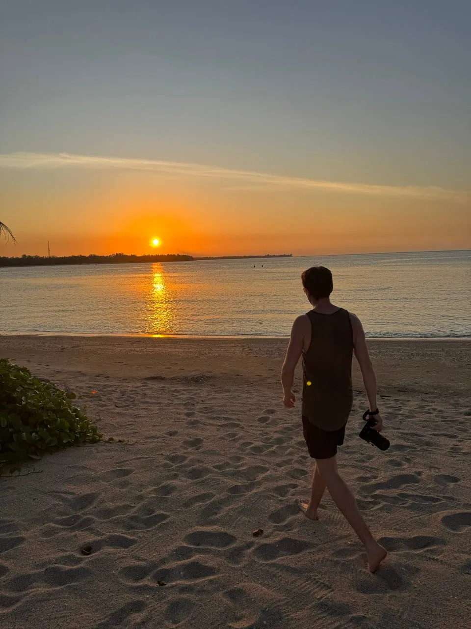 Ein Mann geht barfuss mit einer Kamera in der Hand über den Sandstrand auf die tief stehende Sonne zu