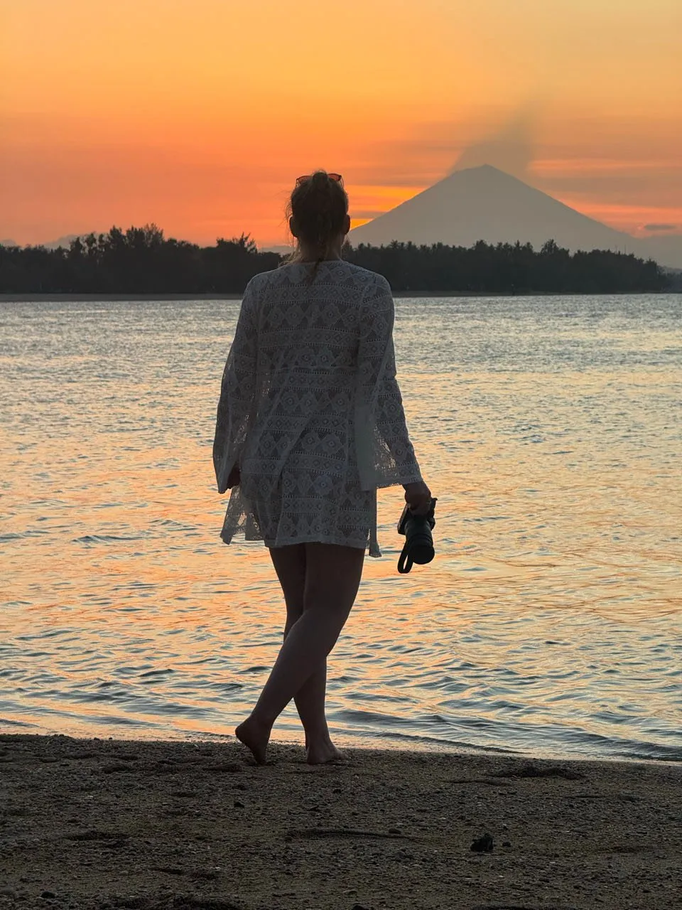 Eine Frau in weissem Spitzenüberwurf steht am Wasser und schaut auf einen Vulkankegel vor orangem Himmel