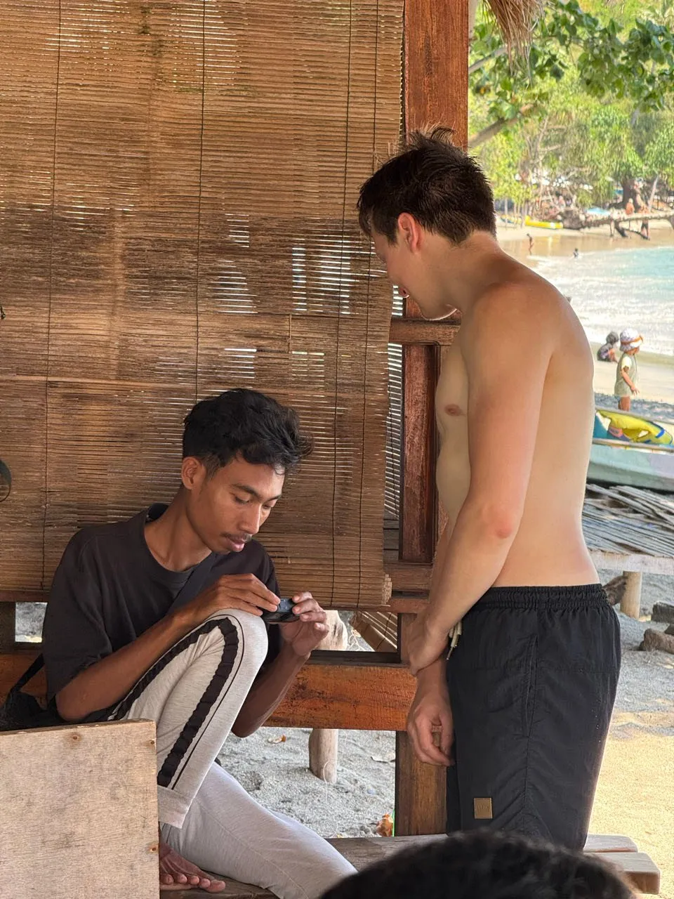 A young local sits in the shade of a hut with a bamboo blind and looks at something in his hands, next to him stands a man in swimming trunks