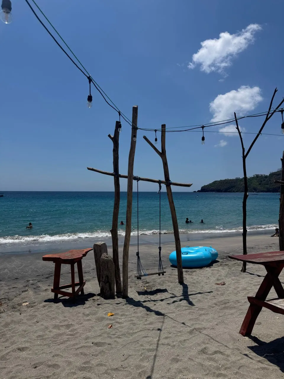 Dark sandy beach with a wooden swing, a blue swim ring and a string of lights, behind it blue sea with surf