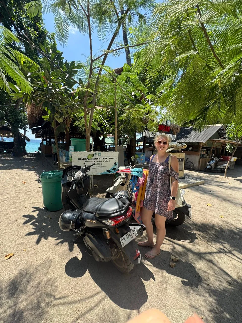 Annina in a patterned dress and sunglasses stands in the shade of trees next to parked scooters, beach huts behind her