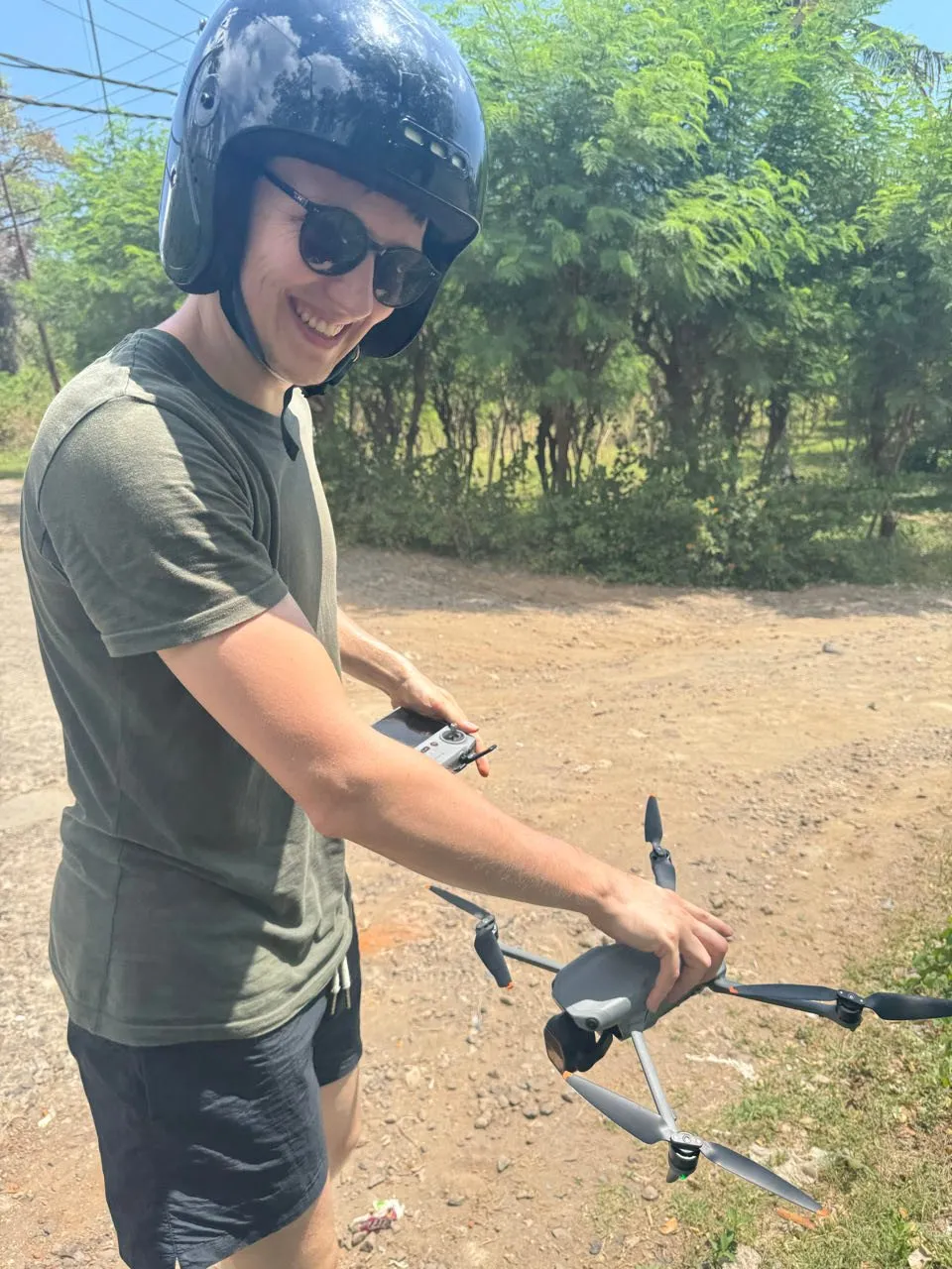 A smiling man with a helmet and sunglasses holds a drone and its remote control in his hands on a dirt track