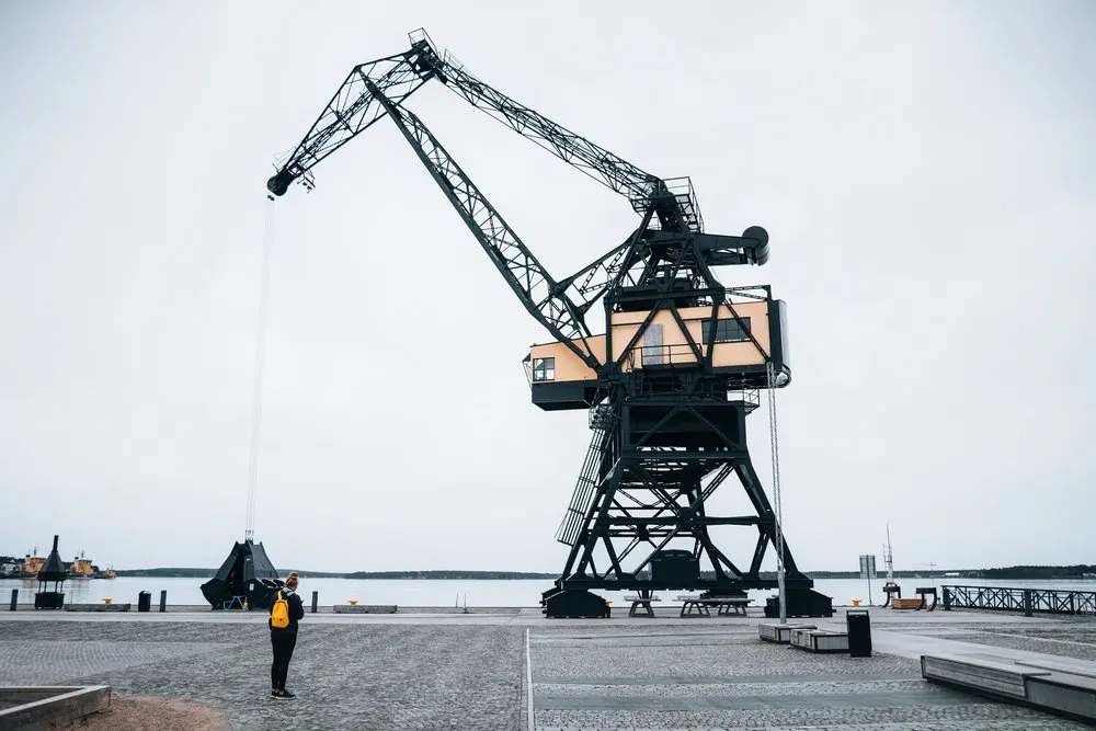 Der schwarze Hafenkran mit gelber Kabine am alten Hafen von Luleå, davor steht eine Person mit gelbem Rucksack