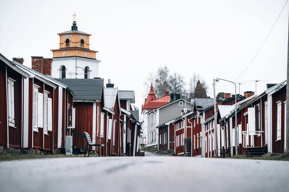 Rote Holzhäuser an einer leeren Strasse in Luleå Gammelstad, dahinter ein weiss und gelb gestrichener Turm
