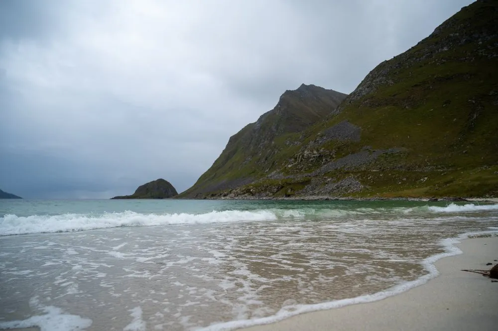 Am Hauklandstranda laufen Wellen auf den Sand, rechts steigt ein steiler grüner Berghang an