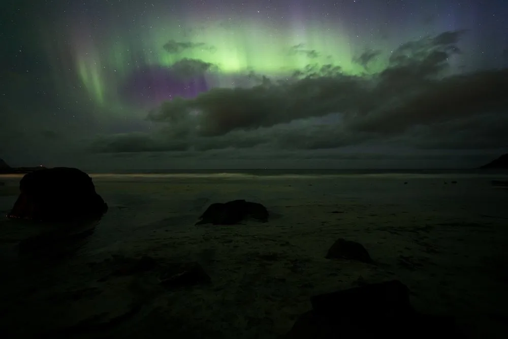 Grüne und violette Nordlichter über dunklen Wolken, darunter ein Strand mit Felsbrocken