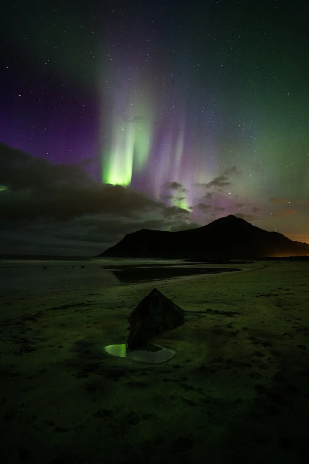Violette und grüne Lichtstreifen über einem dunklen Berg, im Vordergrund ein Fels und eine grün spiegelnde Pfütze im Sand