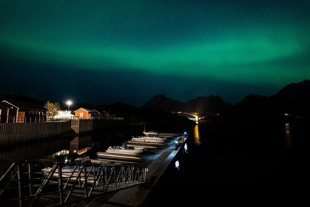 Grünes Nordlicht über einem kleinen Hafen mit Booten am Steg, roten Häusern und einer Brücke im Hintergrund