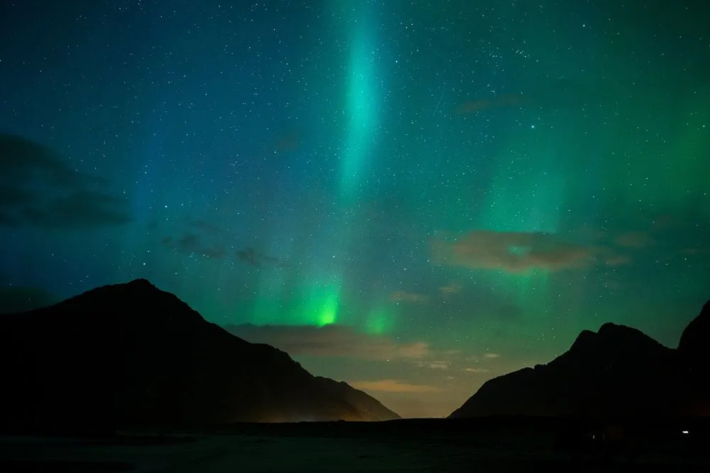 Nordlicht zwischen zwei dunklen Berghängen, am Horizont ein oranger Lichtschein