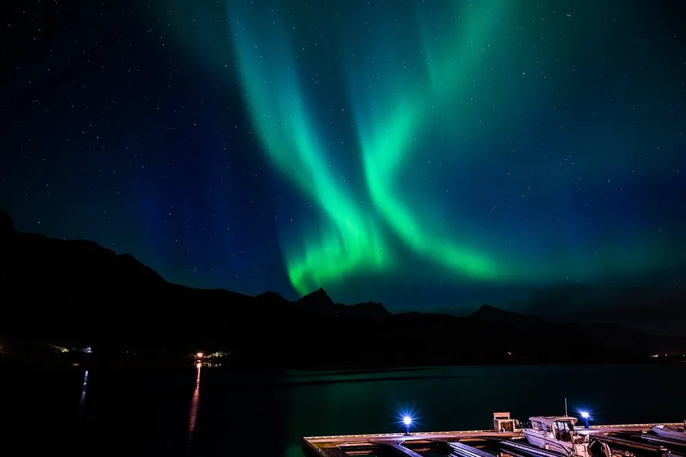 Grüne Lichtbänder über dem Wasser, unten rechts ein Steg mit Booten und blauen Lampen