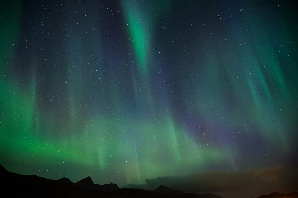 Senkrechte Lichtstreifen in Grün und Violett vor vielen Sternen über einer Bergkette