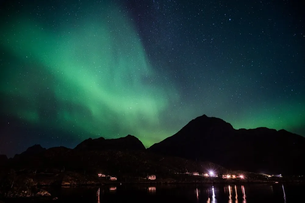 Breite grüne Lichtschleier ziehen schräg über einen Berg, am Ufer brennen Lichter