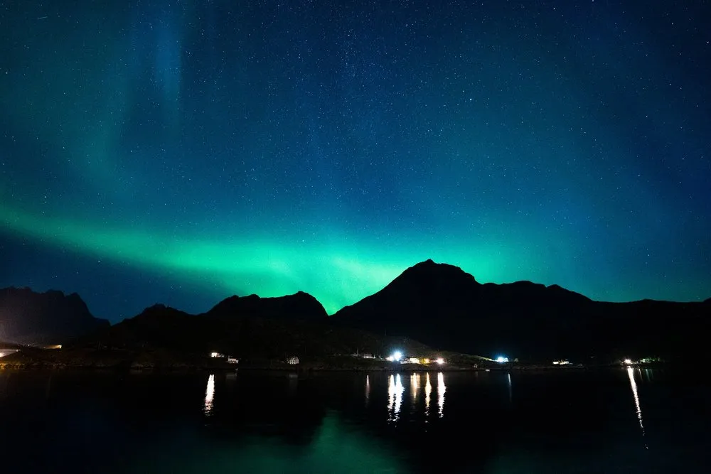 Ein grünes Lichtband hinter einem Berg, die Lichter am Ufer spiegeln sich im ruhigen Wasser