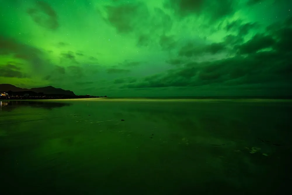 Der ganze Himmel leuchtet grün, darunter ein nasser Sandstrand, der das Licht spiegelt