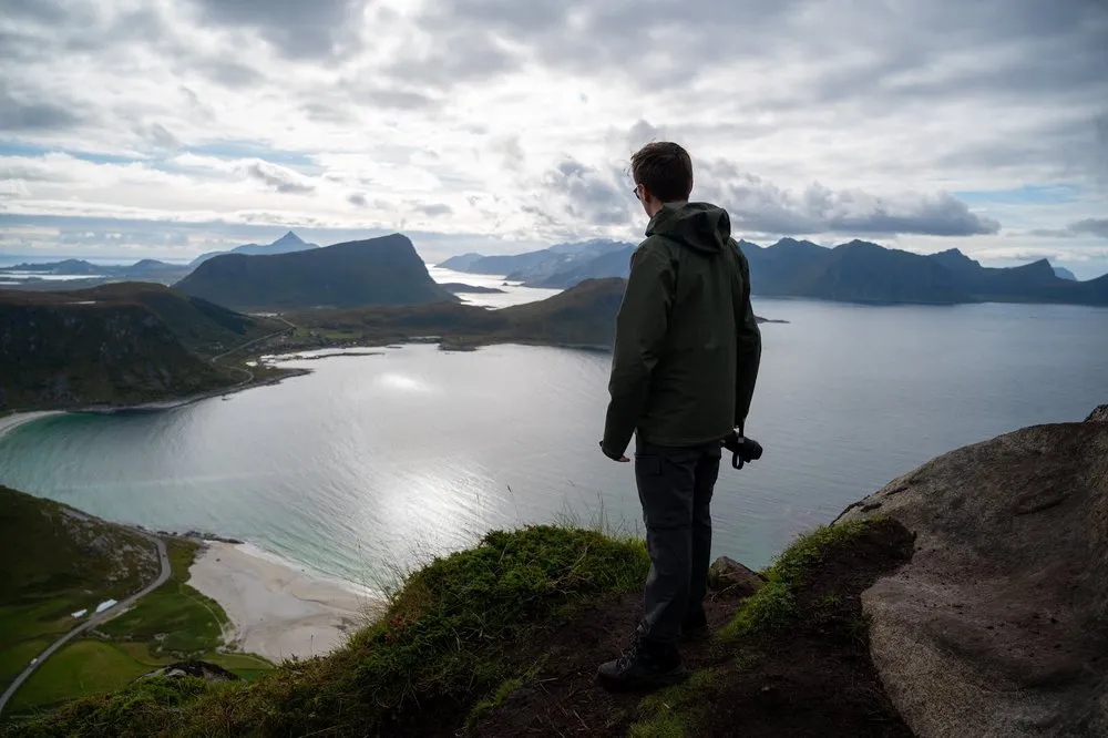 Ein Mann in grüner Jacke steht mit der Kamera in der Hand am Rand und schaut auf Bucht und Berge