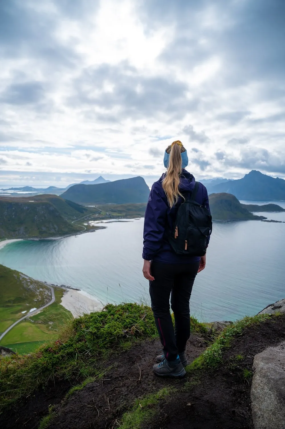 Eine Frau mit Rucksack und blauem Stirnband steht am Rand und schaut auf Bucht und Berge