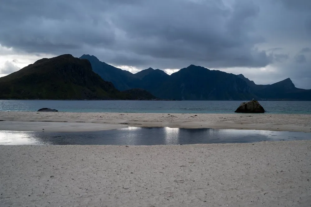 Am Hauklandstranda spiegelt eine Pfütze im hellen Sand den Himmel, dahinter ein Felsblock und dunkle Berge