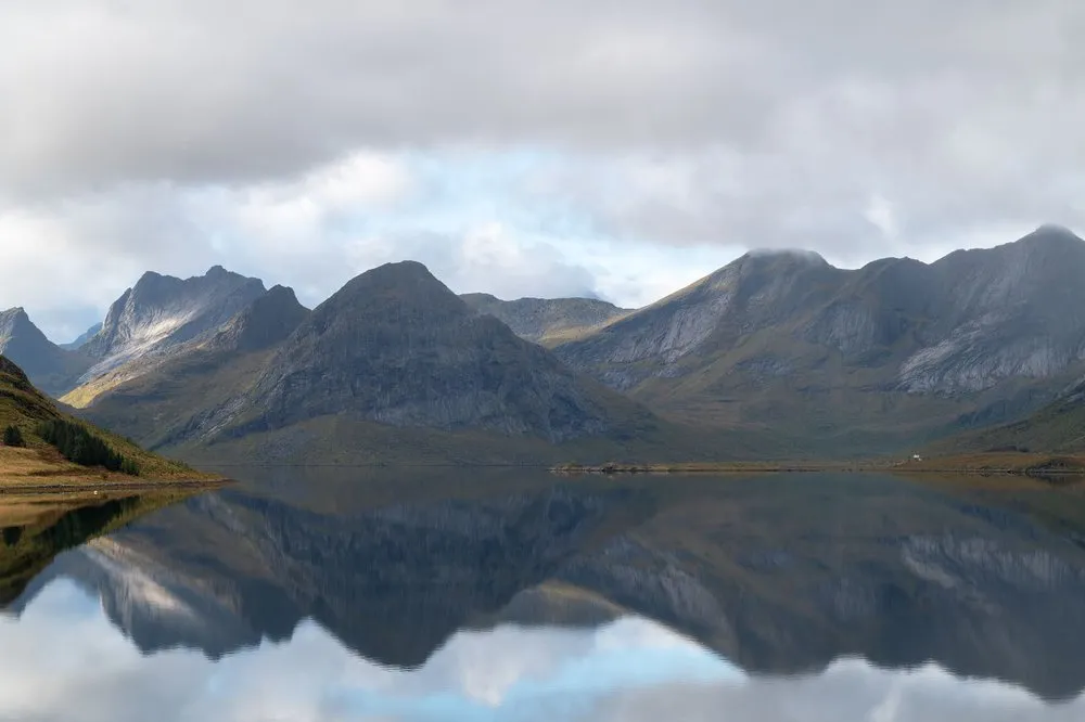 Eine Bergkette spiegelt sich im spiegelglatten Wasser, darüber Wolken mit blauen Lücken