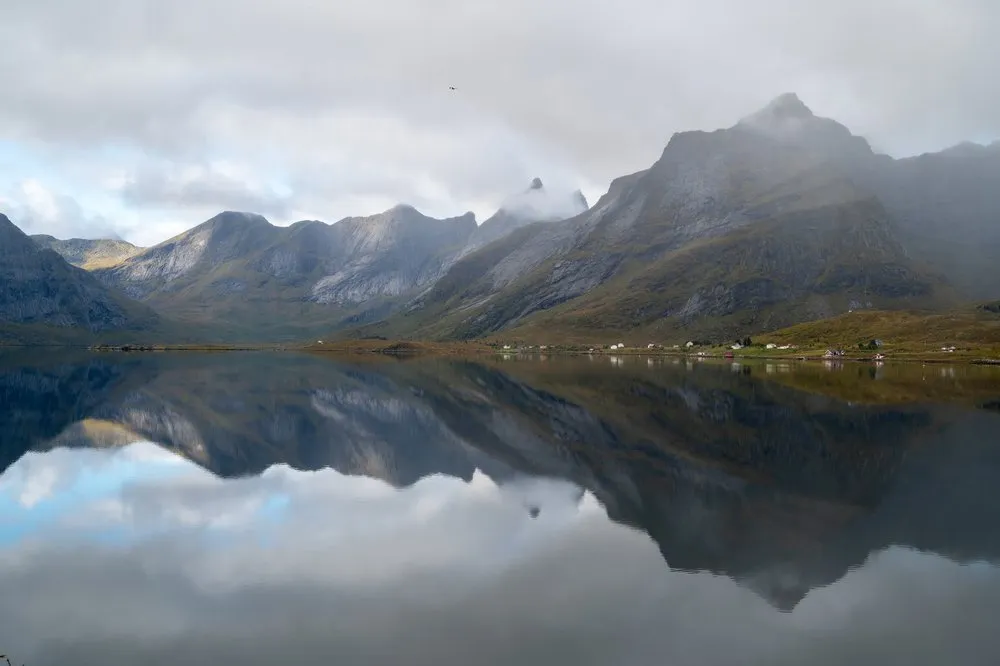 Eine Bergkette unter Wolken spiegelt sich im glatten Meer, am Ufer stehen verstreute Häuser