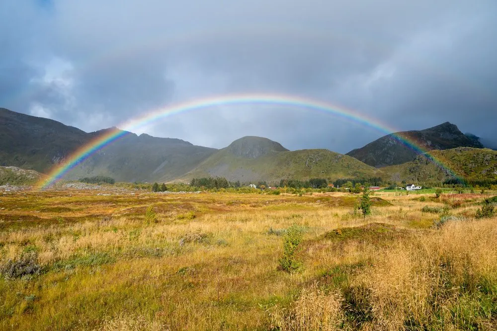 Ein Regenbogen wölbt sich über eine gelbe Herbstwiese und dunkle Hügel, rechts ein weisses Haus