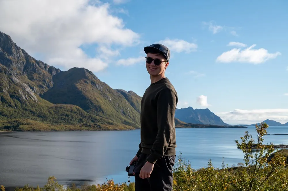 Ein Mann mit Cap und Sonnenbrille hält eine Kamera in der Hand, hinter ihm Fjord, Berge und blauer Himmel