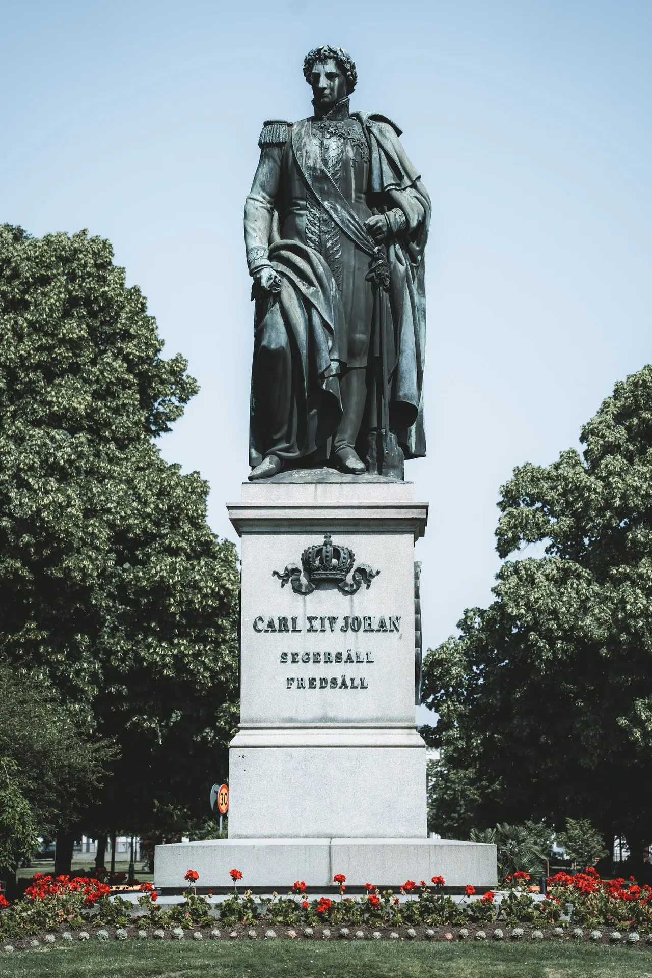 Bronze statue with the inscription Carl XIV Johan on a stone plinth, surrounded by trees and red flowers