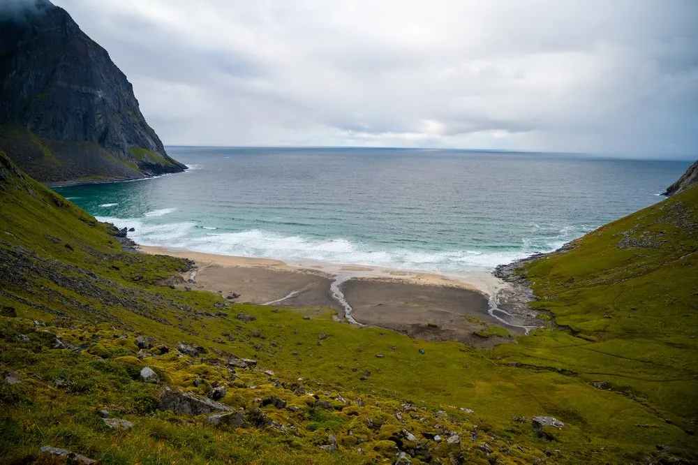 Blick von oben auf die Kvalvika Beach: heller Sand mit Bachlauf, Brandung und grüne Hänge zu beiden Seiten