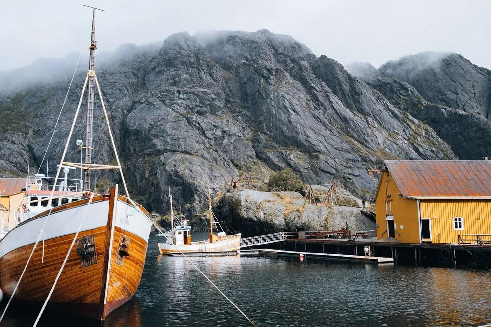 Ein hölzernes Segelboot liegt im kleinen Hafen neben einem gelben Holzhaus, dahinter steigt eine graue Felswand in den Nebel