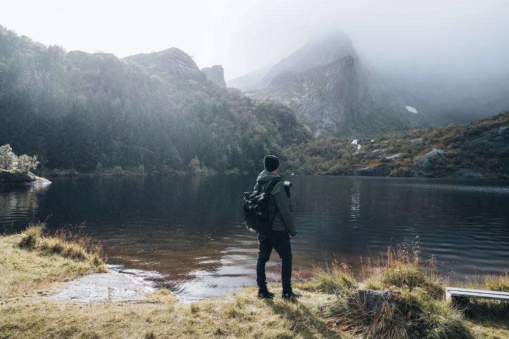 A person with a backpack stands on the shore of a mountain lake, mountains rising in the fog behind