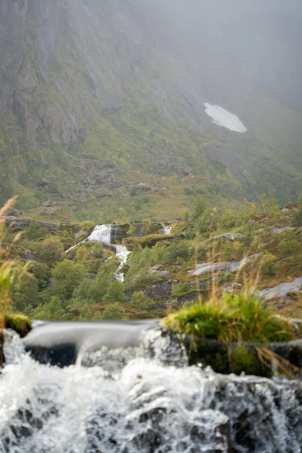A stream tumbles over rocky steps, behind it a mountain slope with small waterfalls and a snowfield in the fog