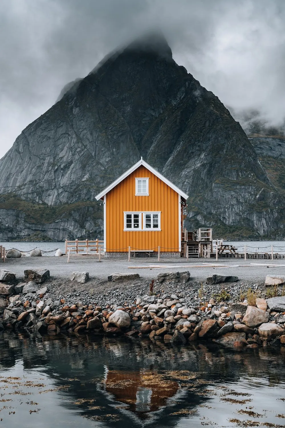 The yellow wooden house in Sakrisøy in front of a steep, fog-shrouded mountain, stones and reflecting water in front of it