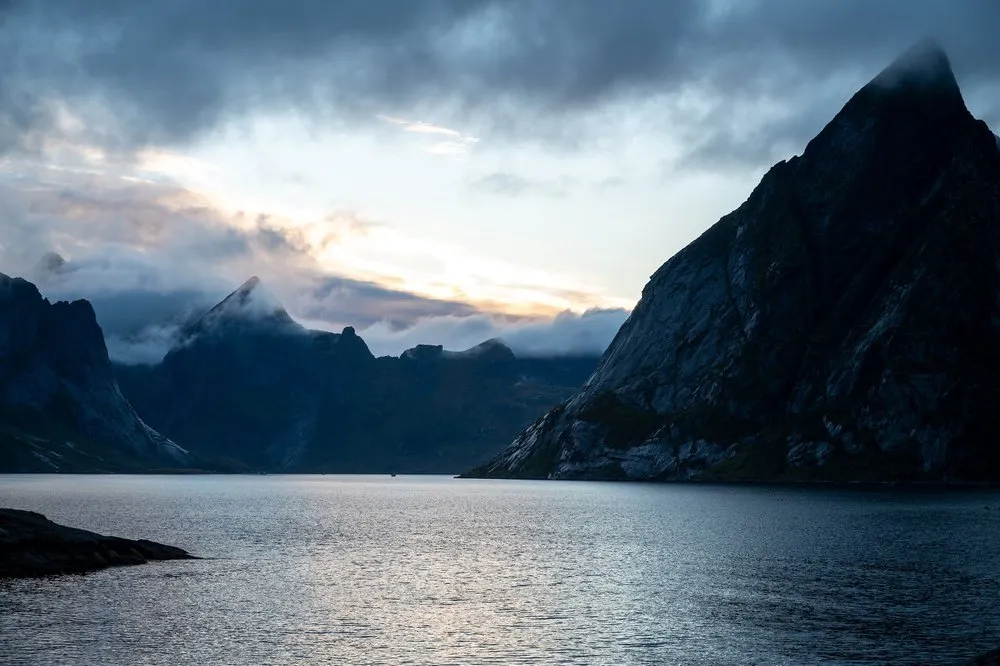 Evening light breaks through clouds above a fjord, a dark mountain peak rising on the right