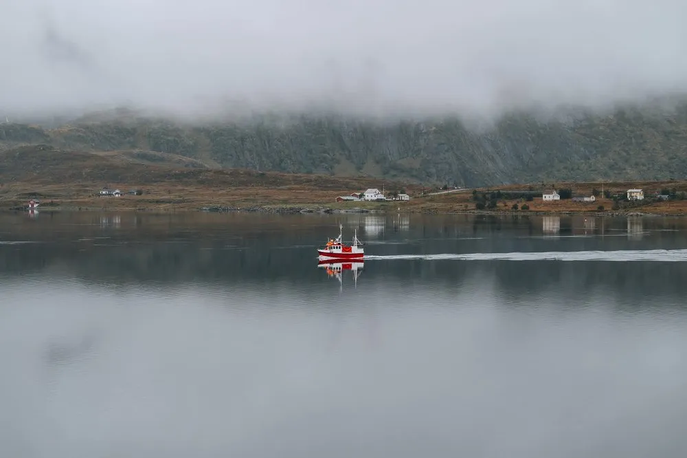 A red boat with a white wake moves across mirror-smooth water, a few houses on the shore under low clouds