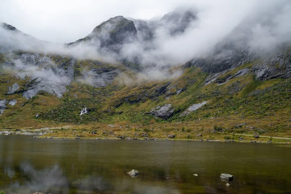 Fog drifts over a mountain slope with small waterfalls, a dark lake with stones in front of it