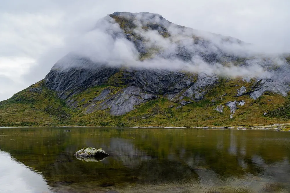 A mountain with a band of cloud at its summit is reflected in the lake, a mossy stone in the foreground