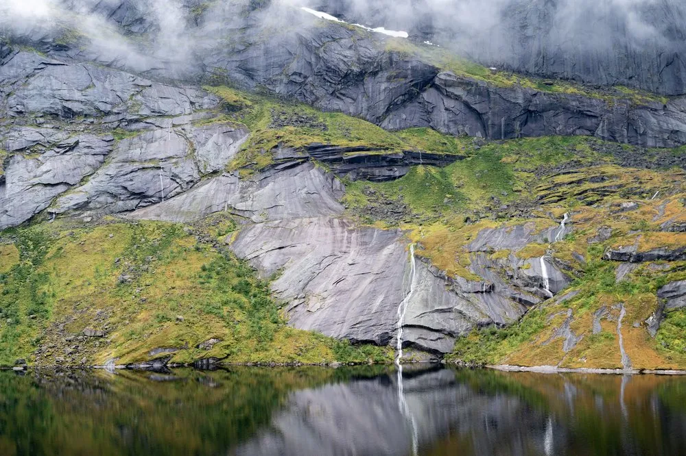 A grey rock face with green and yellow patches, several narrow waterfalls falling down it, is reflected in the lake