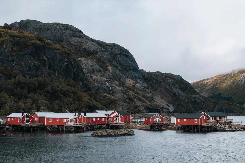 A row of red rorbuer on stilts at the water in Nusfjord, a dark rock slope behind them
