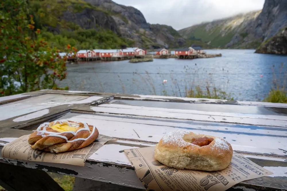 Two pastries on newspaper on an old wooden table, behind them the water of Nusfjord with red rorbuer
