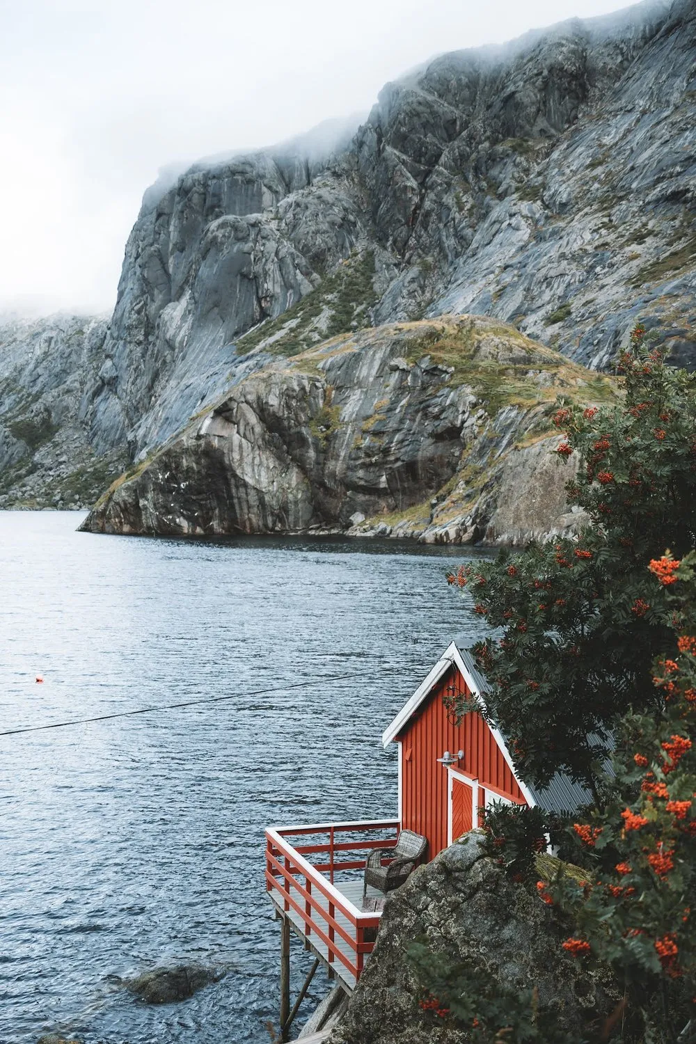 A red wooden cabin with a terrace on stilts stands above the water in front of a steep rock face, beside it a tree with orange berries