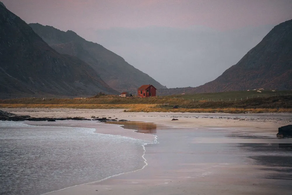 Ein kleines rotes Häuschen hinter dem Flakstadstranda, davor die flachen Wellen auf dem Sand im Abendlicht