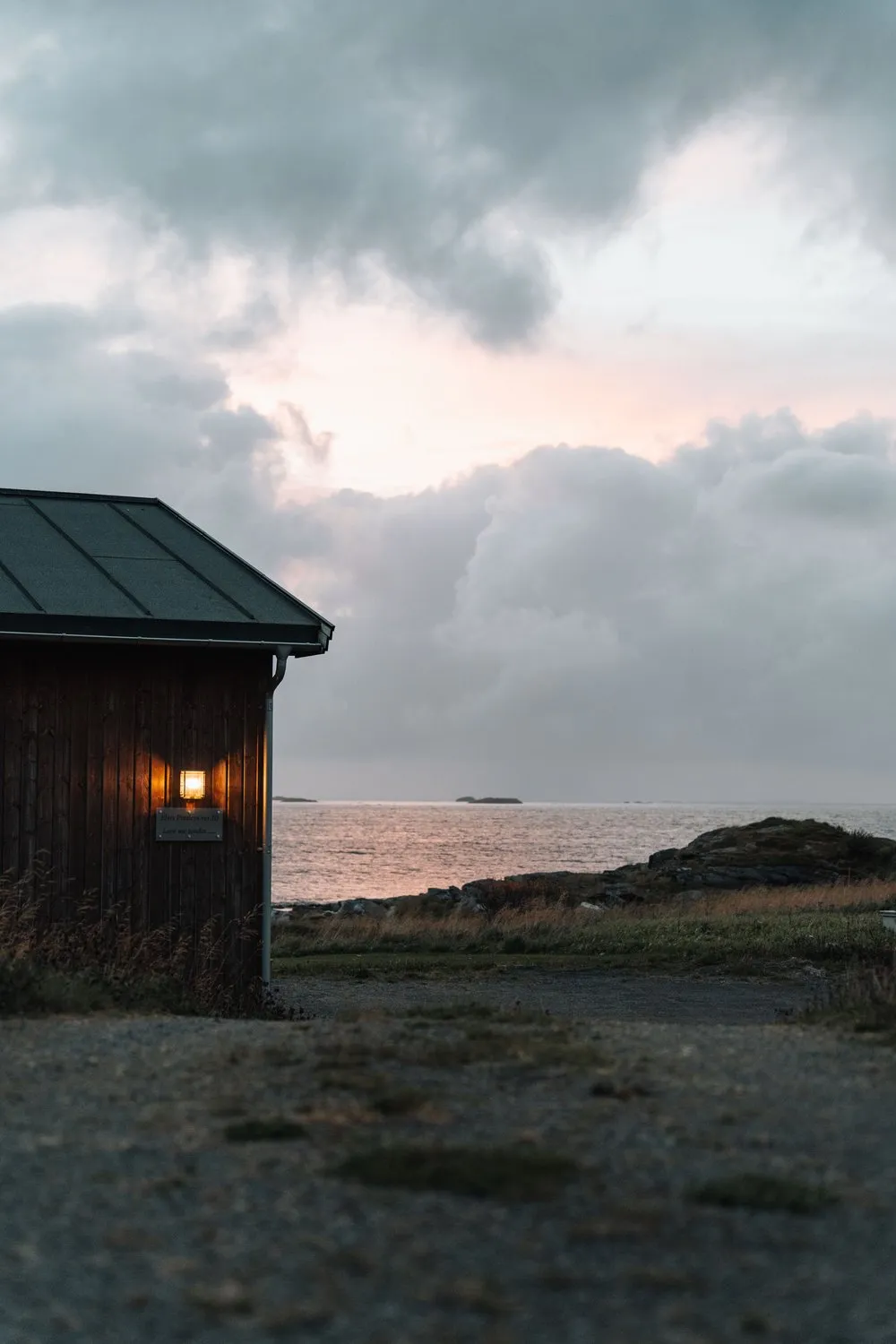 Eine dunkle Holzhütte mit brennender Lampe in der Abenddämmerung, dahinter das Meer unter rosa Wolken