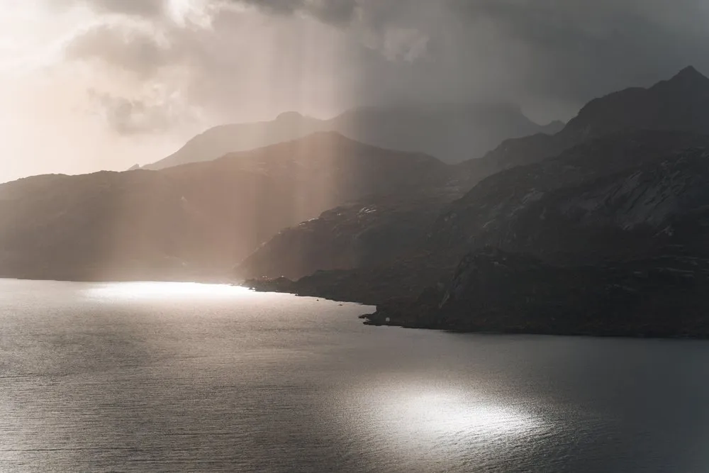 Sonnenstrahlen fallen beim Aufstieg zum Offersøykammen durch die Wolken auf das Wasser vor den dunklen Bergen
