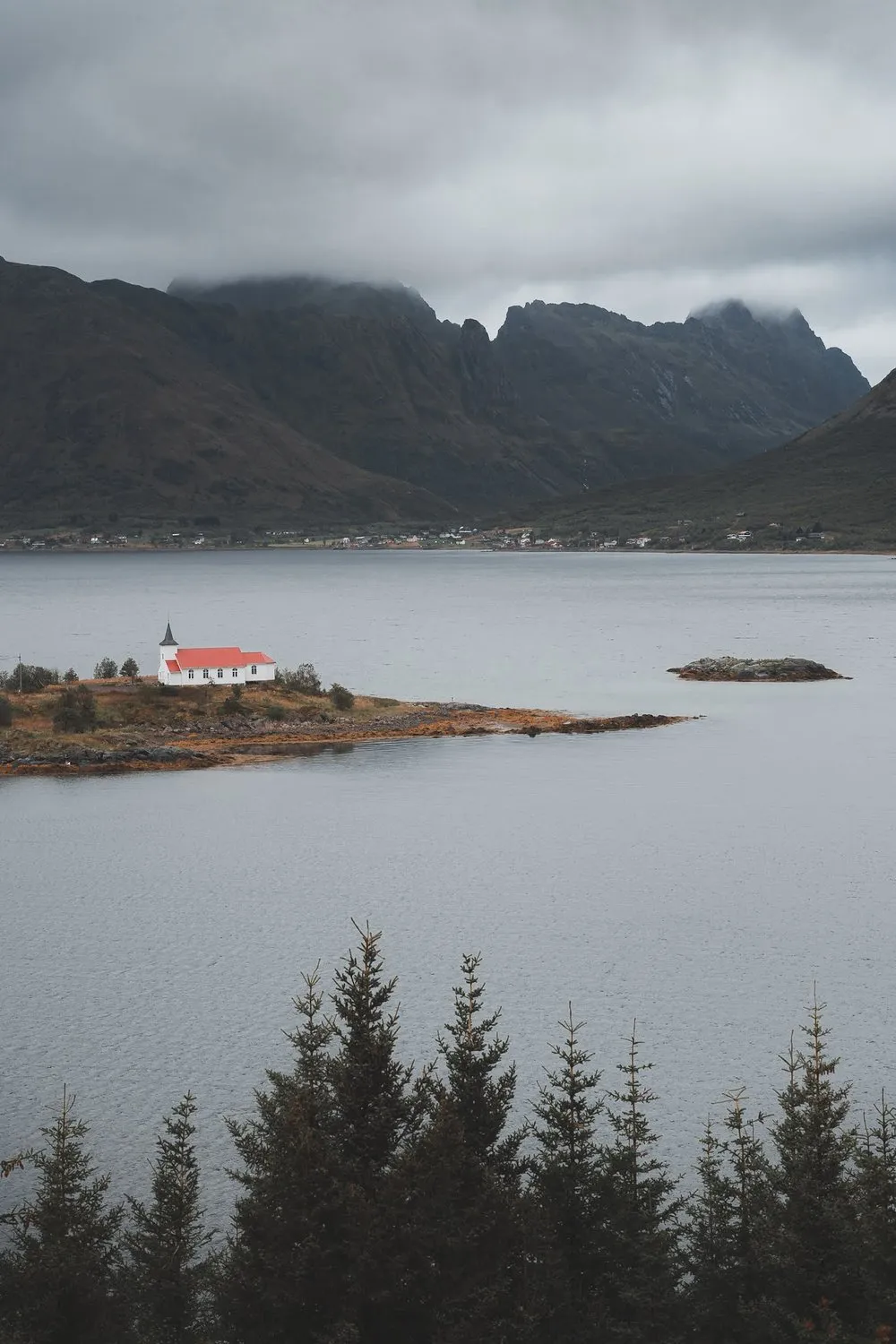 Kleine weisse Kirche mit rotem Dach auf einer Landzunge im Wasser, dahinter Berge in dunklen Wolken
