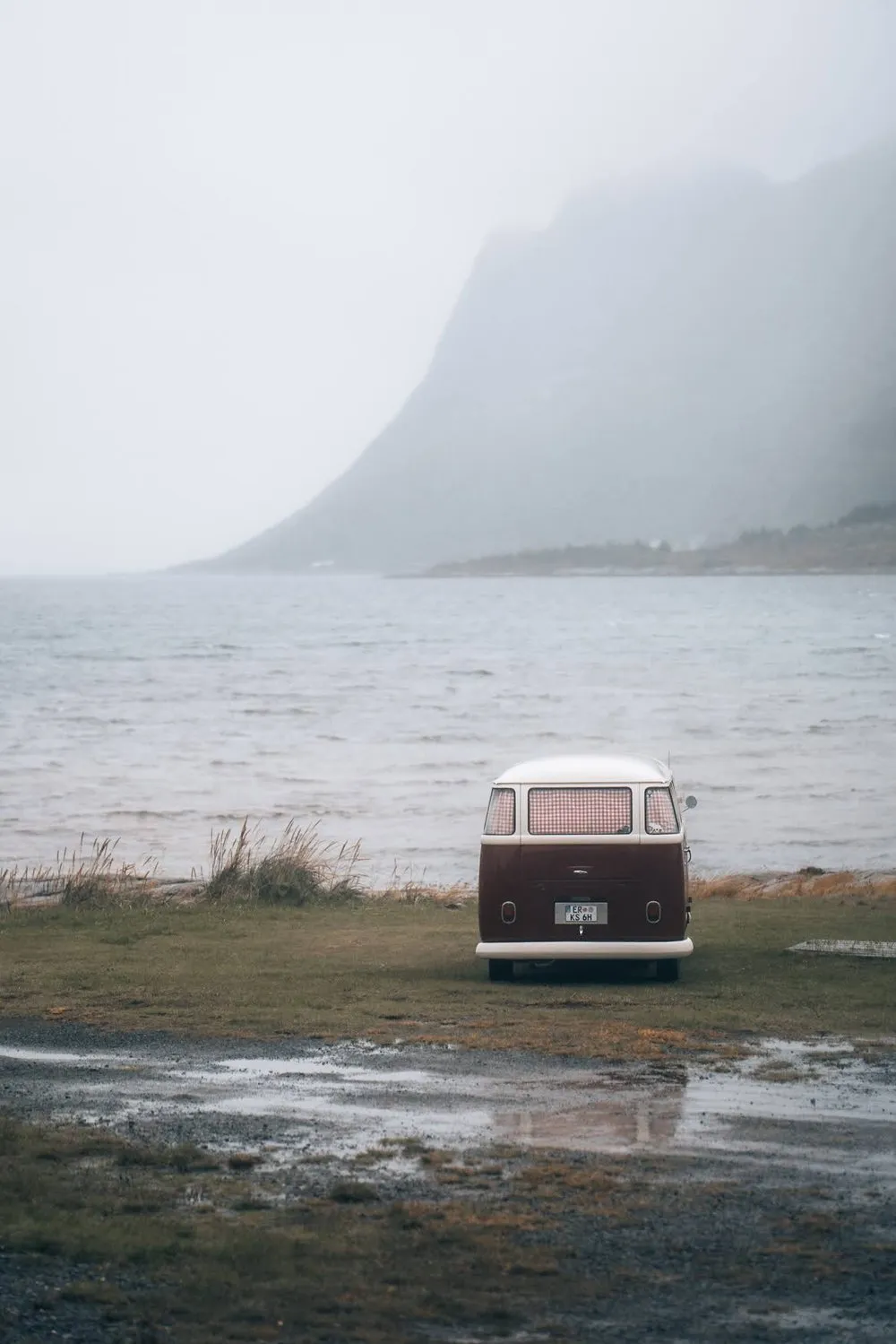Ein alter VW-Bus steht auf einer Wiese am Wasser, dahinter Nebel und ein steiler Berghang