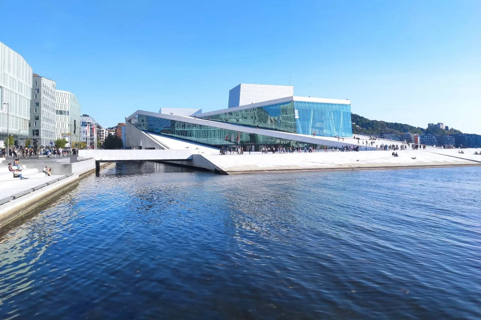 Das Opernhaus mit Glasfassade und schrägen weissen Flächen direkt am Wasser, auf den Rampen sitzen und gehen Menschen