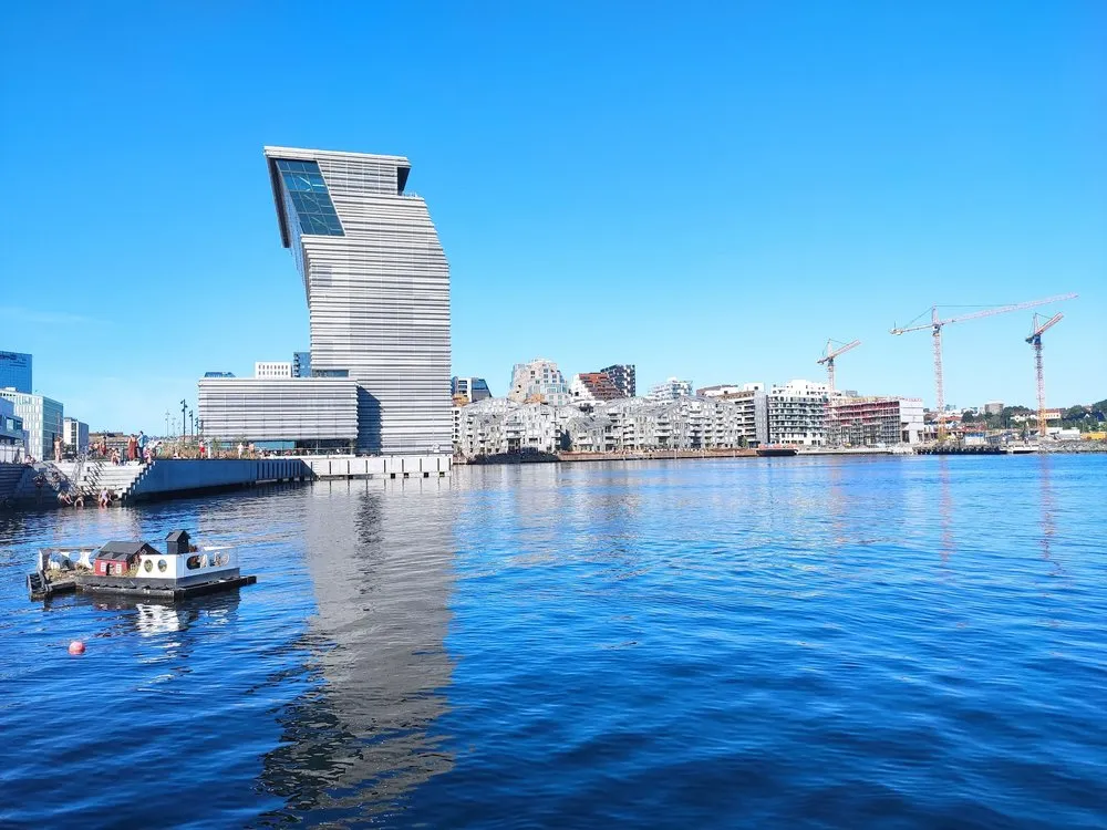 Ein schmaler grauer Turm mit schräg abgeschnittenem Dach am Hafenbecken, links ein kleines Hausboot, rechts Neubauten und Baukräne