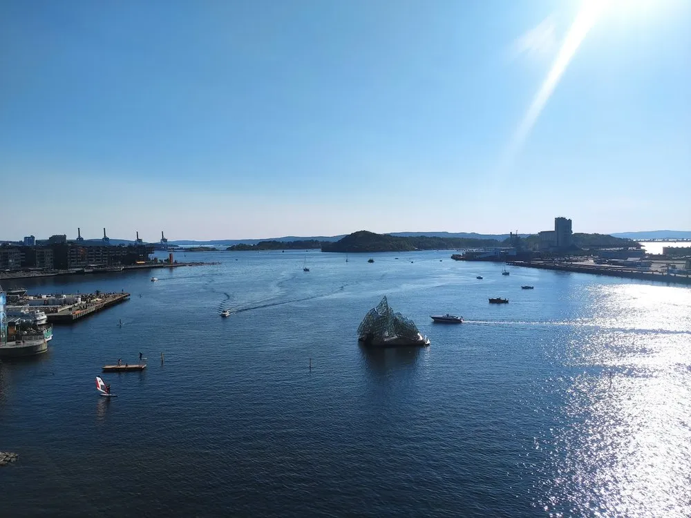 Blick von oben über das Wasser mit Booten und einer Glasskulptur, am Horizont bewaldete Inseln, rechts Sonnenglitzern
